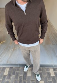 Brauner Reißverschluss-Pullover über einem hellblauen Hemd, kombiniert mit beigen Hosen und weißen Sneakers mit grünen Akzenten, auf einem gefliesten Boden.