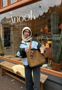 Stribet grøn og blå oversized sweater med en cremefarvet krave, brun ruskinds totebag med en legetøjscharme, sorte jeans, holder en kaffekop.