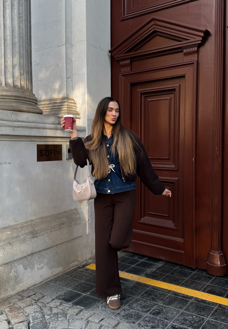 Jeune femme aux longs cheveux tenant une tasse, posant avec une jambe levée à côté d'une grande porte en bois marron et d'une façade de bâtiment en pierre.
