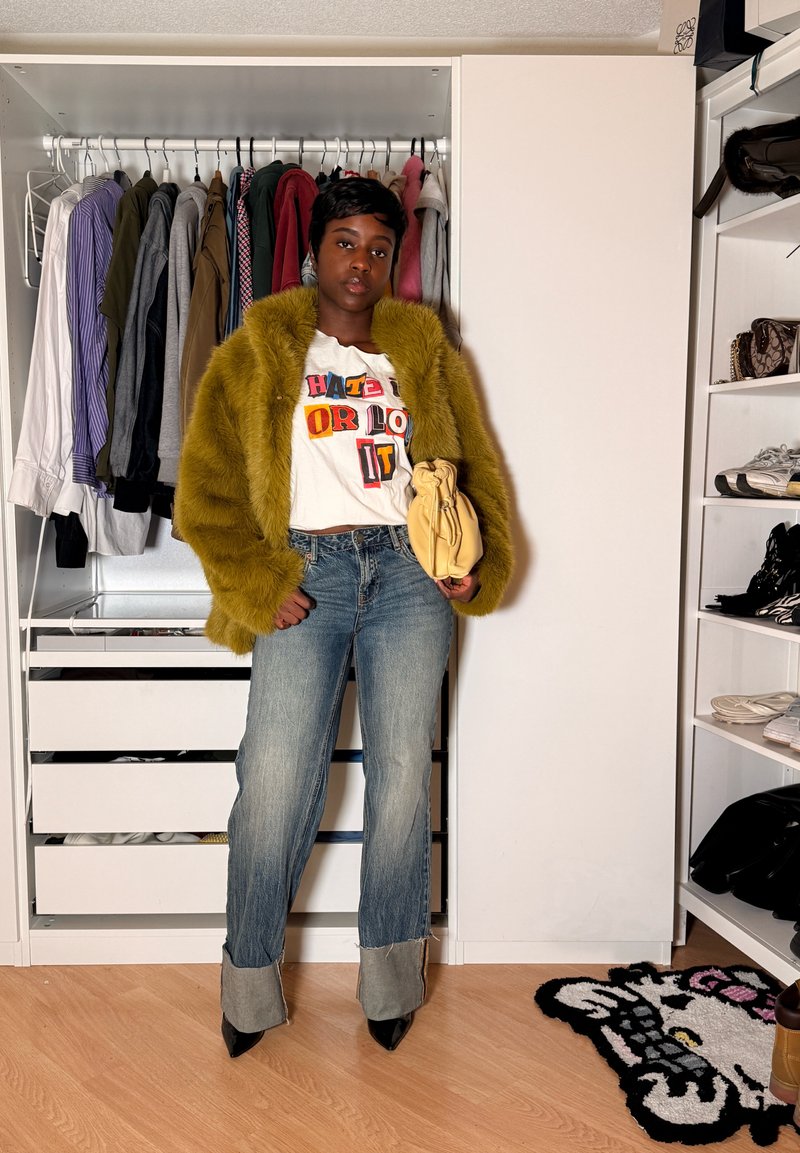 Personne portant une veste en fourrure moutarde, un t-shirt graphique, un jean retroussé et des talons noirs, tenant un sac à main beige, debout dans un placard avec des vêtements et des chaussures.