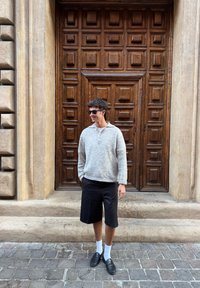 Pulover de lână gri deschis cu guler, pantaloni scurți negri, șosete albe până la gleznă și mocasini negri, stând lângă o ușă texturată din lemn.