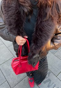 Chaqueta bomber de cuero marrón con cuello de piel sintética, sosteniendo un bolso rojo brillante. Vestido negro con lunares blancos y calzado rojo.