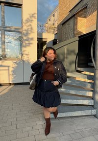 Mujer con chaqueta de mezclilla oscura y falda, con botas marrones, descansando la mano en el cuello, de pie cerca de una escalera de metal en espiral fuera de edificios modernos.