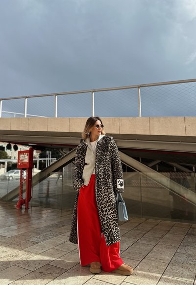 Mujer con abrigo de estampado de leopardo, pantalones anchos rojos, sudadera con capucha beige y gafas de sol, de pie sobre un pavimento de baldosas bajo un paso elevado moderno.