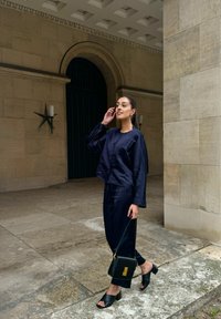 Marinblus av satin med långa ärmar, i kombination med matchande byxor, svart axelväska med guldfärgade detaljer och svarta sandaler.