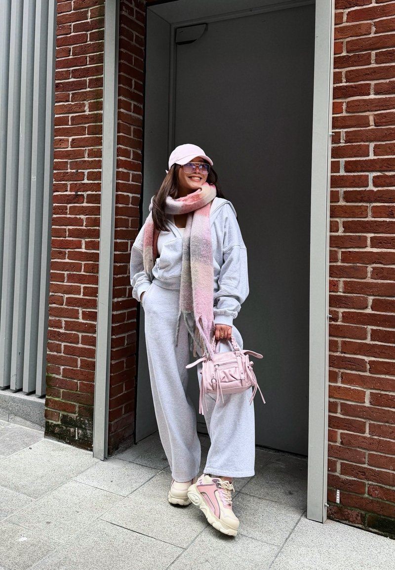 Femme en survêtement gris, écharpe rose, casquette, lunettes de soleil et baskets, souriante, debout à côté d'un mur en briques rouges, tenant un petit sac à main rose.