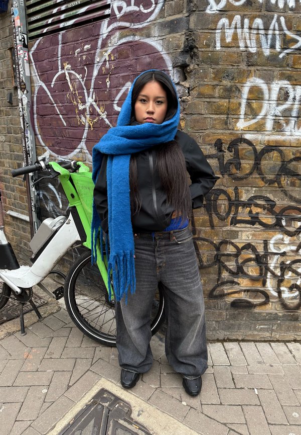 Črna zip-up jakna, modri šal, modra majica, preveliki sivi jeans, črne čevlje; zelena kolo v ozadju, grafiti na opečni steni.
