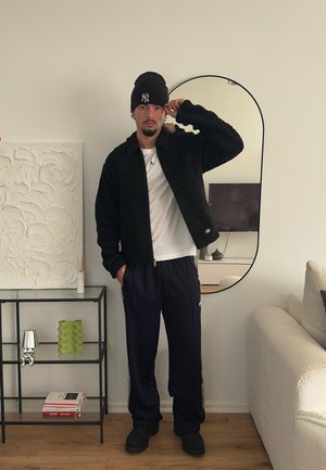 Chaqueta de forro polar negra con cuello, camiseta blanca, pantalones de chándal azul marino con rayas laterales y zapatos negros de slip-on.