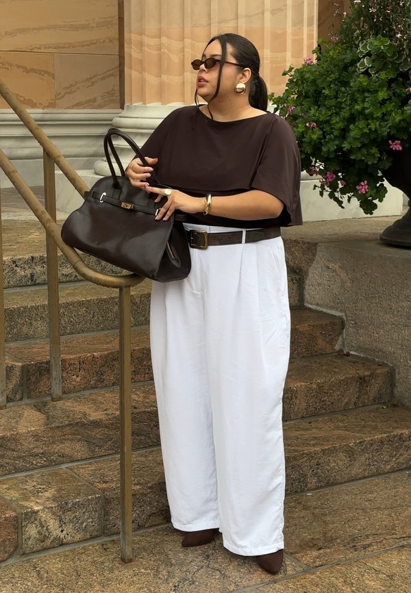 Mujer con camiseta oscura y pantalones blancos está de pie en escalones de piedra sosteniendo un gran bolso negro cerca del pasamanos y una planta con flores rosas.