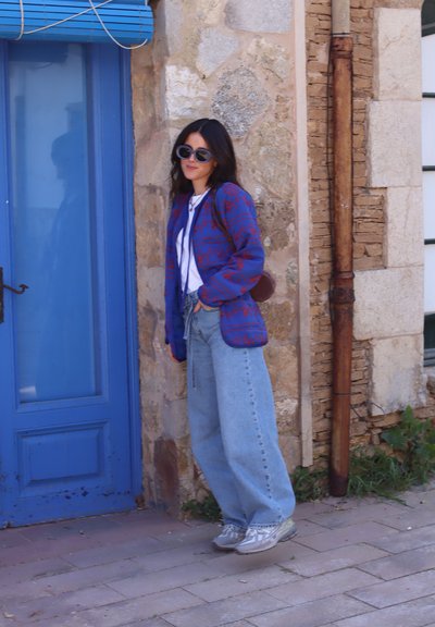 Chaqueta acolchada con un patrón azul y rojo sobre una camisa blanca, combinada con un pantalón vaquero azul suelto y zapatillas grises, frente a una pared de piedra y una puerta azul.