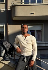 Camisa de manga larga blanca con cuello de muesca, jeans negros y llaves plateadas colgando de un lazo del cinturón, frente a un fondo de edificio gris.