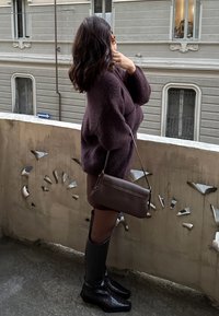 Υπερμεγέθης μωβ πλεκτός σκούφος, καφέ τσάντα ώμου με κομψό σχέδιο, μαύρες καμποτίνα μπότες με υφή, στέκεται σε μπ�αλκόνι.