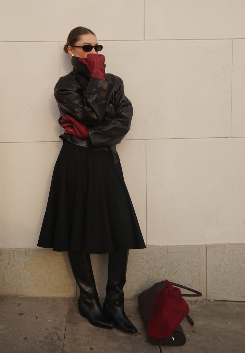 Femme en veste en cuir noire, jupe noire, bottes et gants rouges, appuyée contre un mur ; un sac rouge et marron repose à proximité sur le sol.