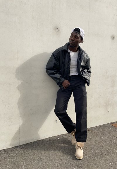 Hombre con gorra blanca al revés, chaqueta de cuero negra, camisa blanca, pantalones negros y botas beige, apoyado contra una pared de hormigón al aire libre.
