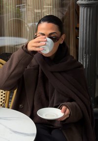 O persoană îmbrăcată într-o vestă maro texturată stă la o masă de marmură, ținând o ceașcă ceramică albă și o farfurioară albă, purtând ochelari de soare.