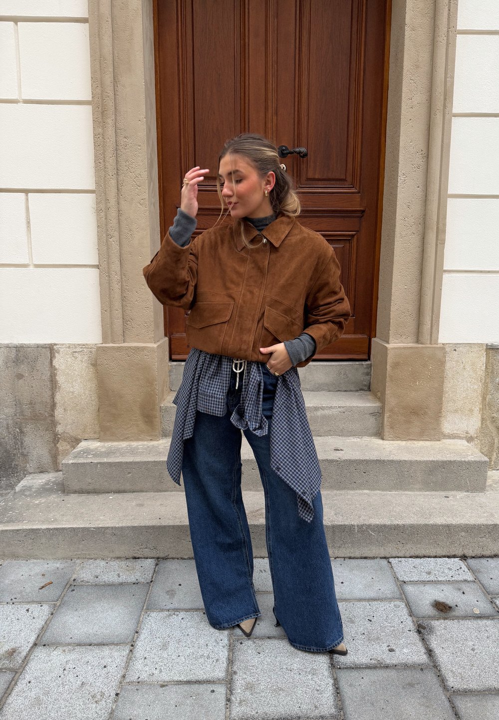 Veste courte en daim marron avec deux poches avant, portée sur un top gris et une chemise à carreaux bleue nouée autour de la taille. Jean en denim à jambes larges.