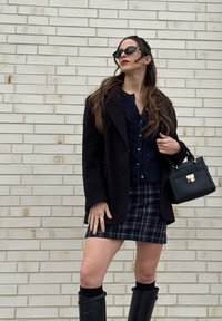 Γυναίκα που φοράει μαύρο παλτό, μπλε πουλόβερ, καρό φούστα, μαύρες μπότες μέχρι το γόνατο, γυαλιά ηλίου και κρατάει μια μαύρη τσάντα χειρός μπροστά σε έναν λευκό τοίχο από τούβλα.