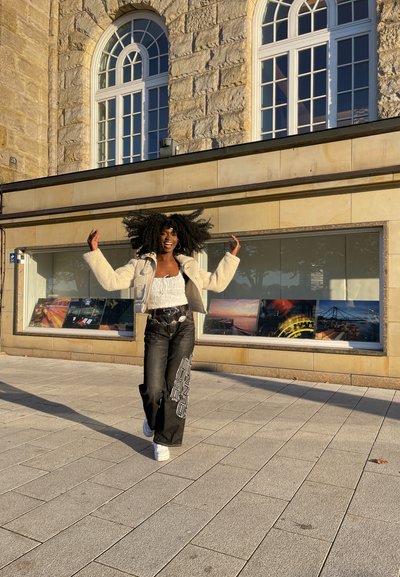 Chaqueta de piel sintética crema sobre una blusa de encaje blanca, combinada con jeans negros que tienen patrones geométricos blancos y zapatillas blancas. Luz brillante del sol de fondo.