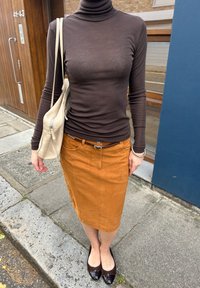 Brun genser med høy hals, lys beige blyantskjørt, svarte leiligheter med detaljer, og en lys beige skulderveske. Glatte teksturer og tettsittende design.