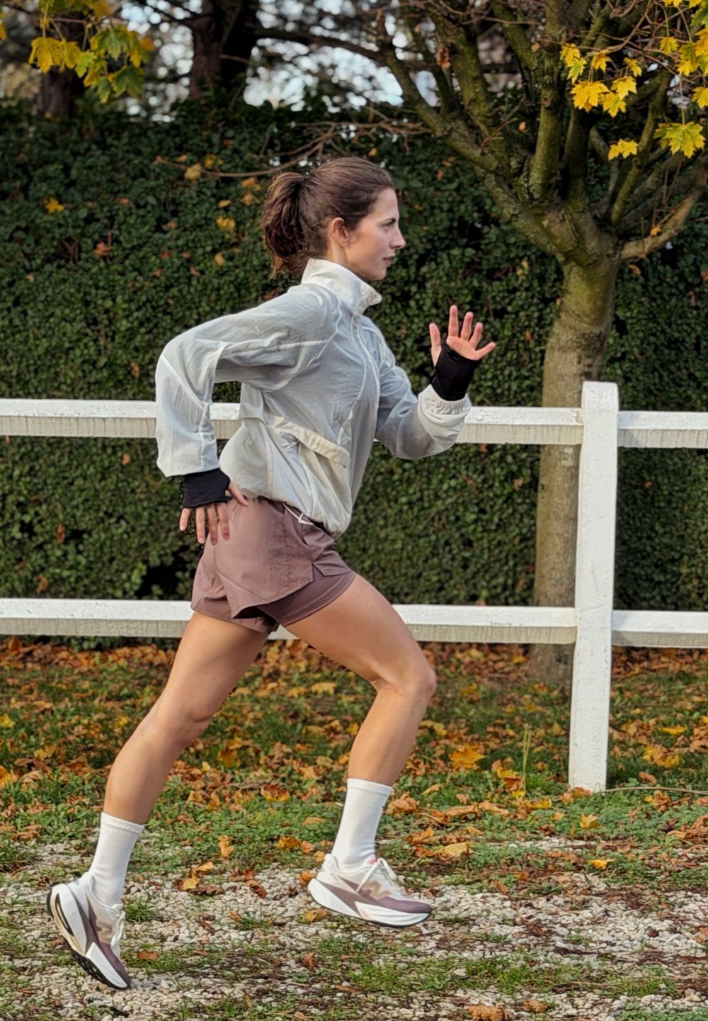 Biegacz w jasnoszarej wiatrówce i brązowych szortach, noszący białe skarpetki i trampki. W tle ogrodzona zieleń z jesiennymi liśćmi.