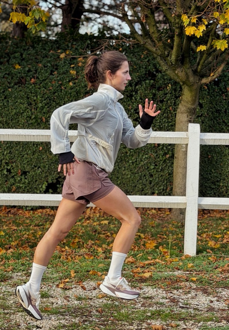 Läufer in einer hellgrauen Windjacke und braunen Shorts, trägt weiße Socken und Turnschuhe. Im Hintergrund ein eingezäuntes Grün mit Herbstblättern.
