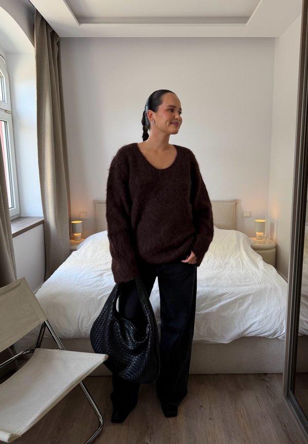 Mujer con suéter y pantalones oscuros sosteniendo una bolsa grande negra, de pie en un dormitorio con una cama blanca y una silla beige cerca de una ventana.