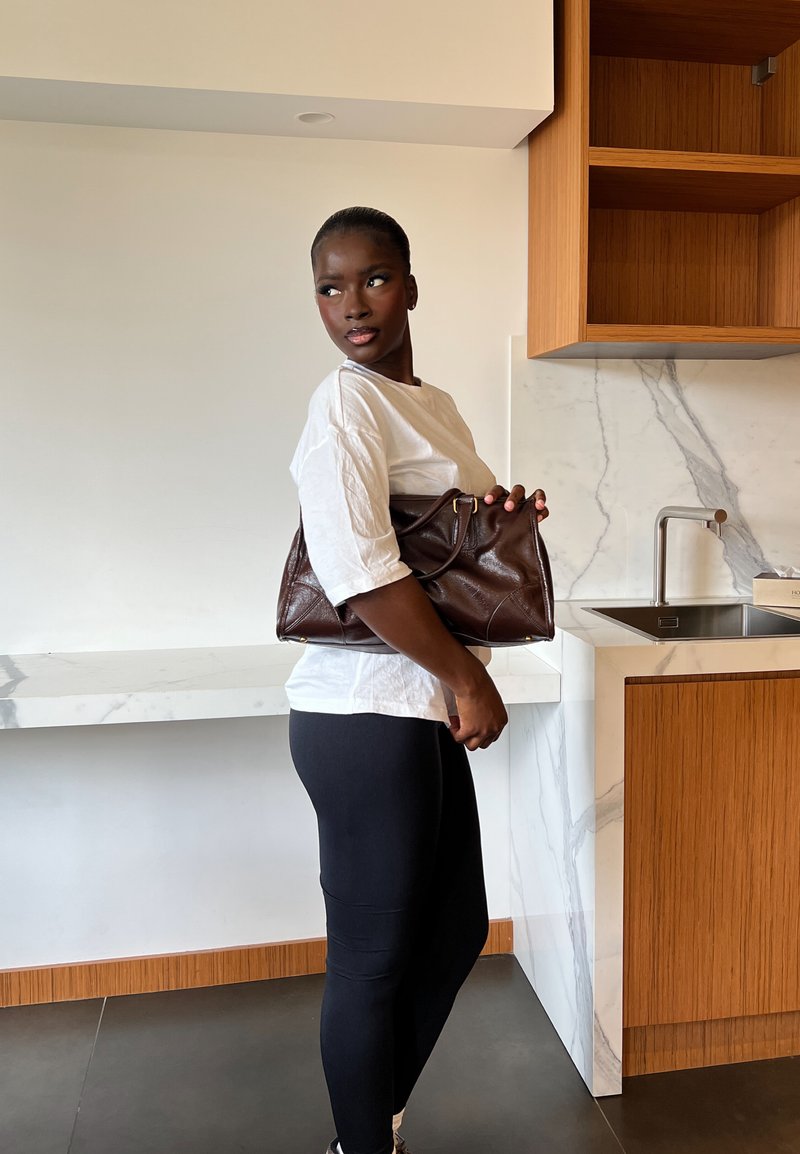 Femme en chemise blanche et leggings noirs tenant un grand sac en cuir marron, debout dans une cuisine moderne avec des plans de travail en marbre et des armoires en bois.