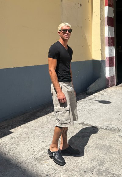 Camiseta negra de cuello en V, pantalones cortos tipo cargo beige con grandes bolsillos, zuecos negros. Fondo claro con sombras sutiles. Entorno urbano minimalista.