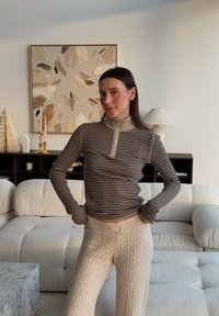 Gestreiftes Langarmoberteil in Beige und Braun mit Reißverschluss am Kragen, kombiniert mit cremefarbenen, grob gestrickten Hosen. Weiche Texturen und figurbetontes Design.