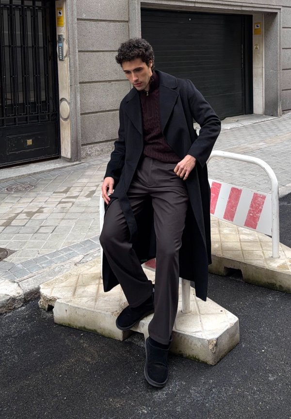 Man wearing long black coat and dark pants sitting on a street barrier in an urban setting with gray paving stones.