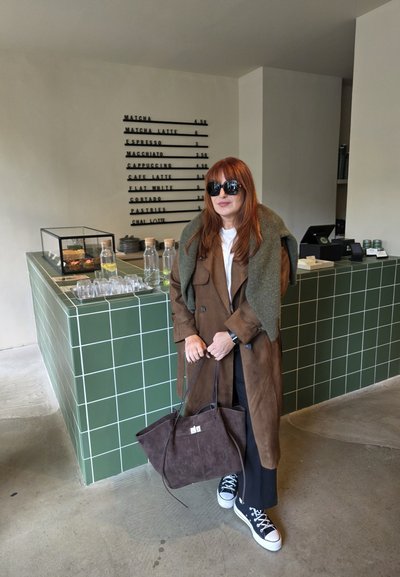 Abrigo de ante marrón, suéter verde oliva, pantalones negros y zapatillas negras con suelas blancas. Sostenido un bolso de ante marrón. Encimera de azulejos verdes al fondo.