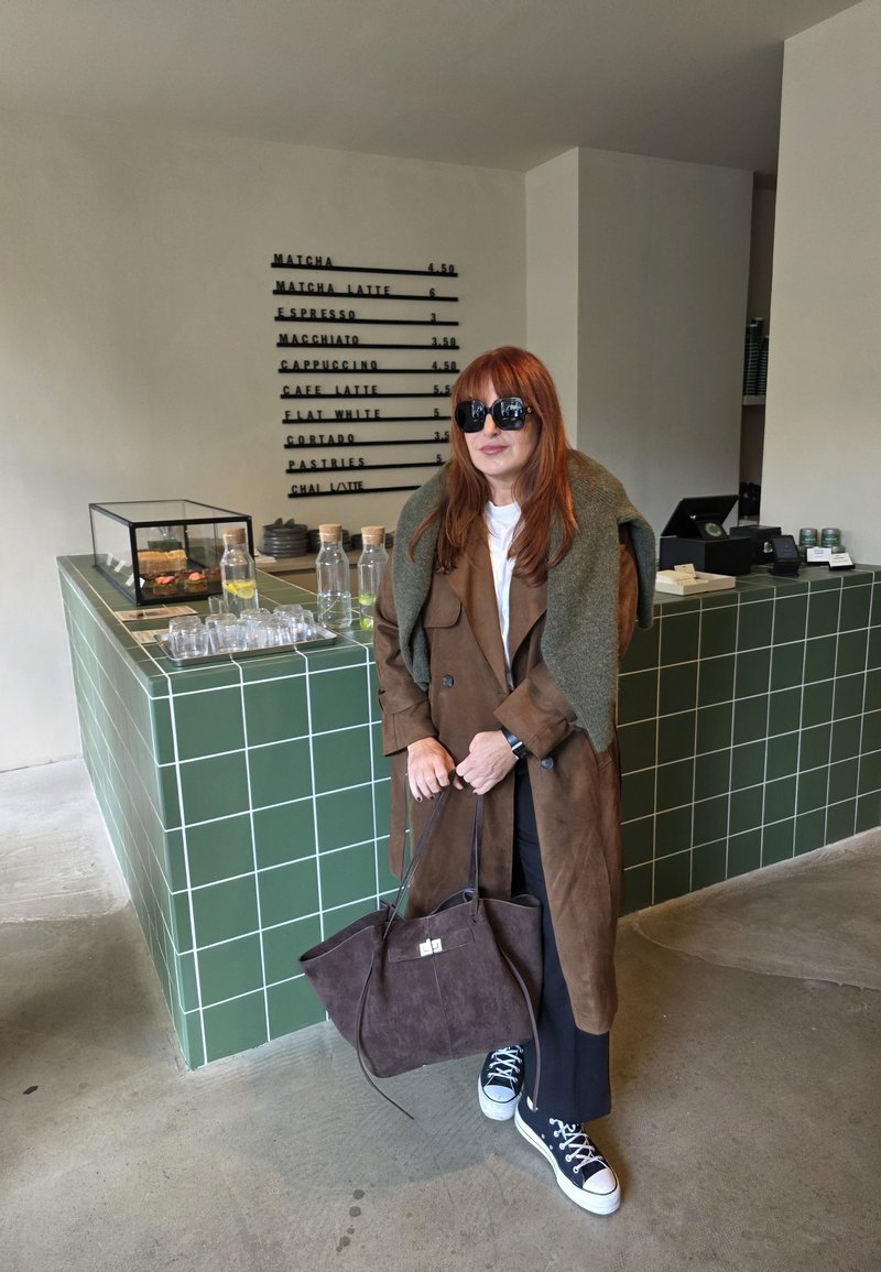Manteau en daim marron, pull vert olive, pantalons noirs et baskets noires avec des semelles blanches. Tient un sac en daim marron. Comptoir carrelé vert en arrière-plan.