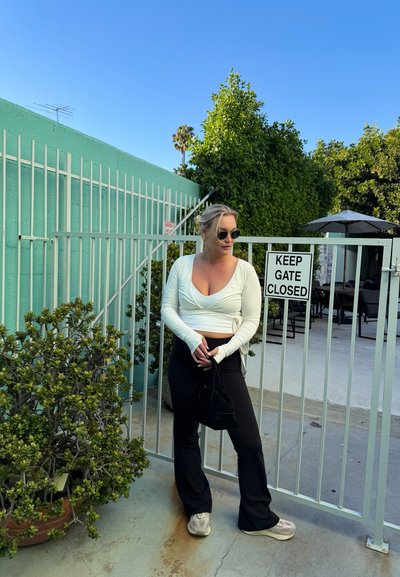 Mujer con gafas de sol, camiseta blanca de manga larga y pantalones negros, de pie frente a una puerta blanca con un cartel que dice "MANTENGA LA PUERTA CERRADA".