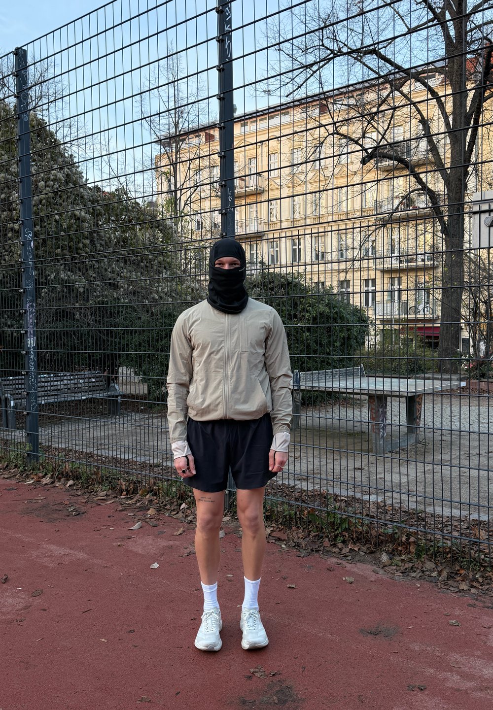 Jachetă bej deschis cu fermoar, mâneci lungi, pantaloni scurți negri, adidași albi și mască de față neagră, stând pe o pistă roșie lângă o gard.