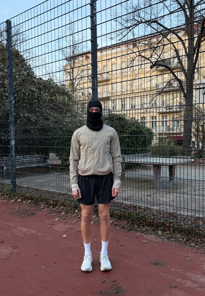 Chaqueta beige claro con cremallera y mangas largas, pantalones cortos negros, zapatillas blancas y mascarilla negra, de pie sobre una pista roja cerca de una valla.