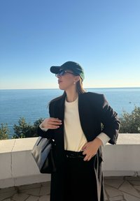 Femme portant un manteau noir et une casquette, un pull blanc, des lunettes de soleil, et portant un sac noir, debout près d'une rambarde avec la mer et un ciel dégagé en arrière-plan.