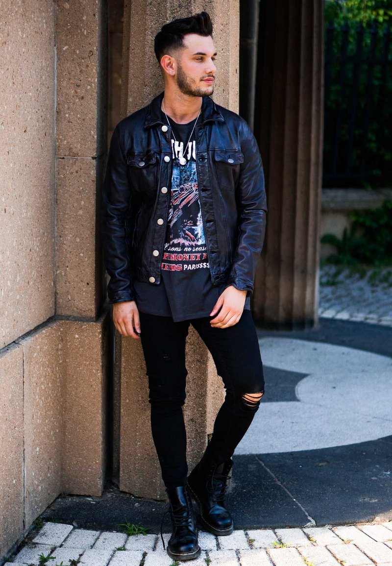 Schwarze Lederjacke über einem bedruckten T-Shirt, kombiniert mit zerrissenen schwarzen Jeans und schwarzen Kampfstiefeln. Der Hintergrund zeigt Steine und Grünpflanzen.