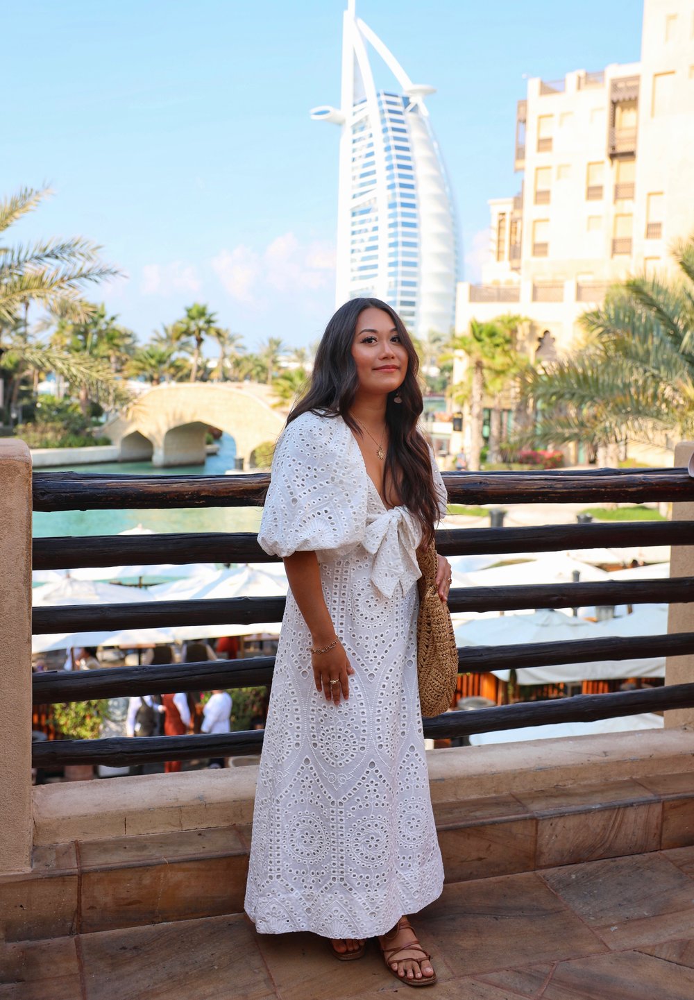 White maxi dress featuring an eyelet pattern, puff sleeves, and a bow at the waist. Paired with a brown woven bag and sandals, against a backdrop of palm trees and buildings.