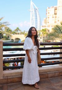 White maxi dress featuring an eyelet pattern, puff sleeves, and a bow at the waist. Paired with a brown woven bag and sandals, against a backdrop of palm trees and buildings.