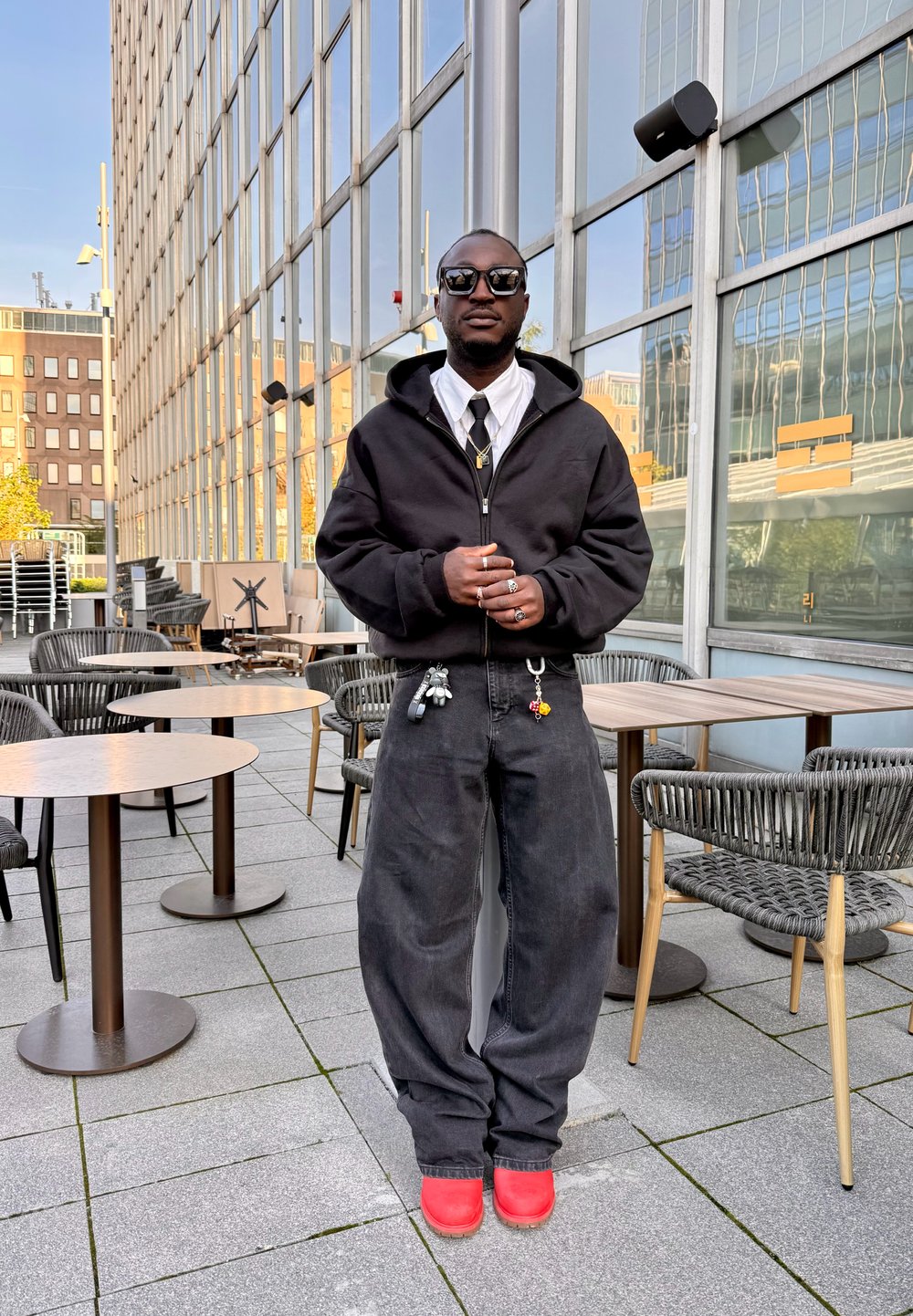 Vīrietis valkā saulesbrilles, melnu kapuci, baltu kreklu, melnas plašas bikses un sarkanas kurpes, stāv ar rokām sakrustotām uz āra terases pie galdiem un krēsliem.