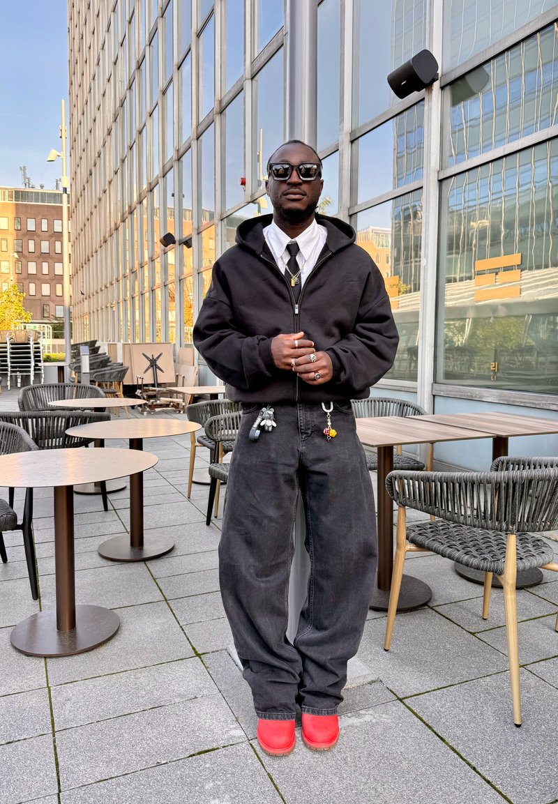 Homme portant des lunettes de soleil, un sweat à capuche noir, une chemise blanche, un pantalon large noir, des chaussures rouges, debout les mains jointes sur une terrasse extérieure avec des tables et des chaises.