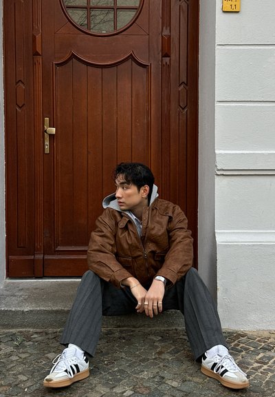 Chaqueta de cuero marrón sobre un hoodie gris, pantalones a rayas grises y zapatillas blancas con rayas negras. Sentado junto a una puerta de madera.