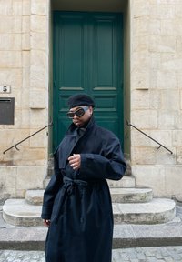 Dolgi črni plašč s pasom, visokim ovratnikom in prevelikimi črnimi sončnimi očali, proti ozadju kamnite stene in zeljenih vrat.