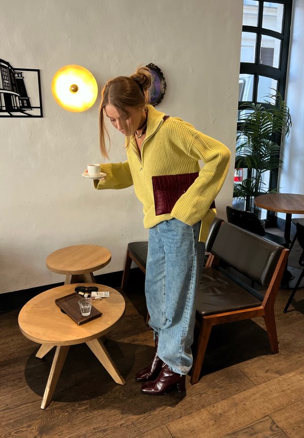 Una mujer con un jersey amarillo y vaqueros sostiene una taza pequeña en una cafetería, junto a mesas y sillas de madera.