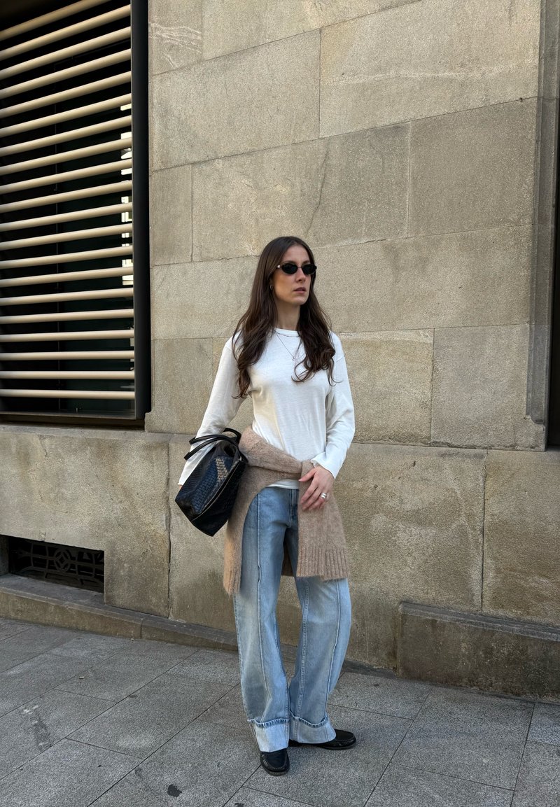 White long-sleeved shirt, light blue wide-leg jeans, beige knit jumper draped, black handbag, and sunglasses against a stone wall backdrop.