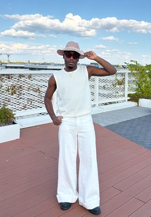 Top sin mangas blanco acanalado, pantalones anchos blancos y sueltos, botas negras hasta el tobillo, y un sombrero de paja beige. Entorno al aire libre con un cielo nublado.