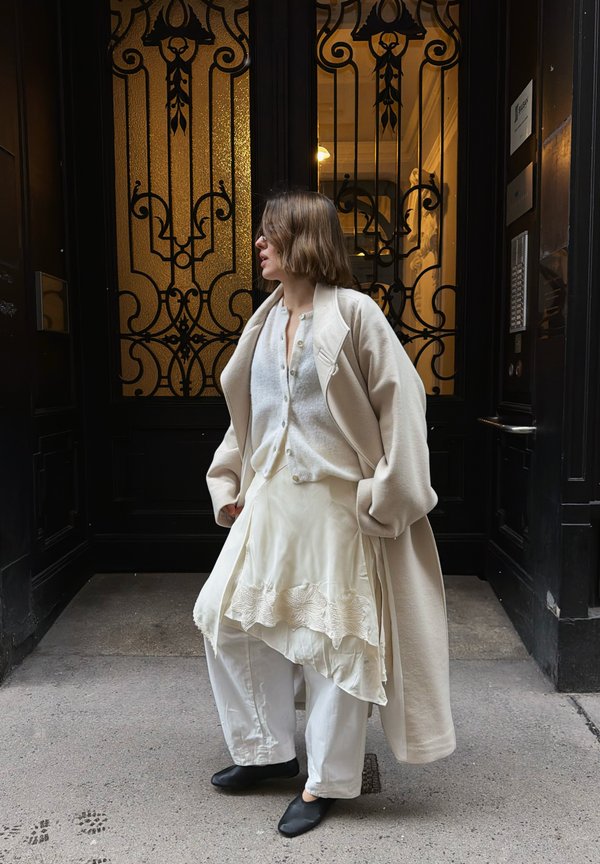 Femme en tenue blanche superposée et manteau beige se tient près de portes noires et dorées ornées, regardant vers la gauche sur un pavé en pierre.