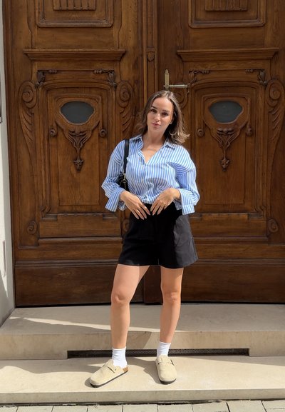 Camisa de botones a rayas azul y blanca, pantalones cortos negros y zapatos de lona beige. De pie frente a una puerta de madera con detalles ornamentales.