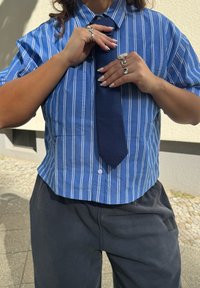 Īsām piedurknēm, zila svītraina krekls ar apkakli, valkāts ar tumšu teksturētu kaklasaiti, papildināts ar sudraba gredzeniem un nagiem.
