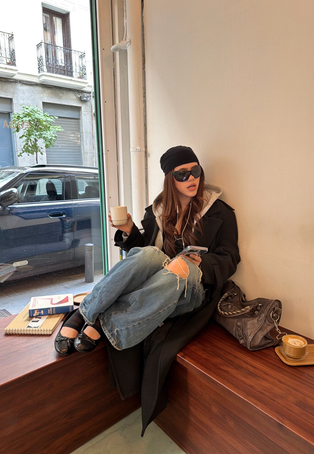 Svart overcoat med grå hettegenser, slitte blå jeans, svarte flate sko, solbriller og en beanie. Holder en kaffekopp, med telefon ved siden av.